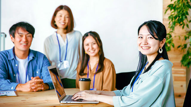 スタッフが笑顔でカメラを見つめる写真。左端の女性はノートPCに手を置き、特に明るく笑っています。私たちのチームの連帯感と、お客様やユーザーへの情熱を反映しています。